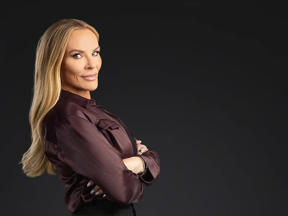 Heather Gay with folded arms in a burgundy blouse against a black background.