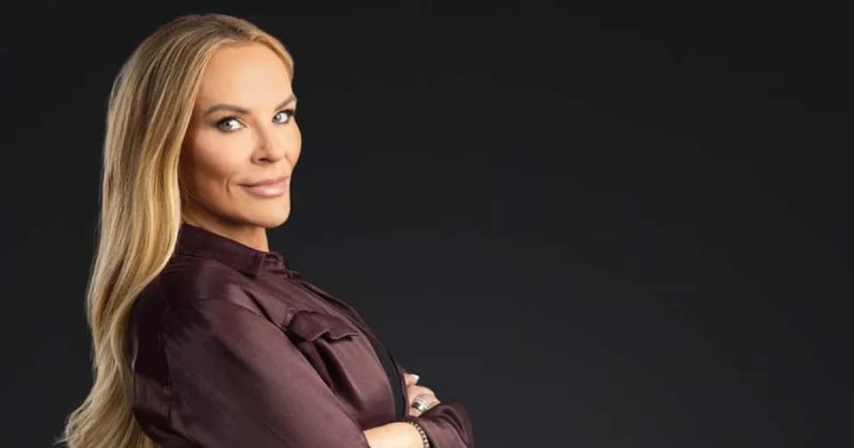 Heather Gay in a burgundy blouse with her arms folded, photographed against black.